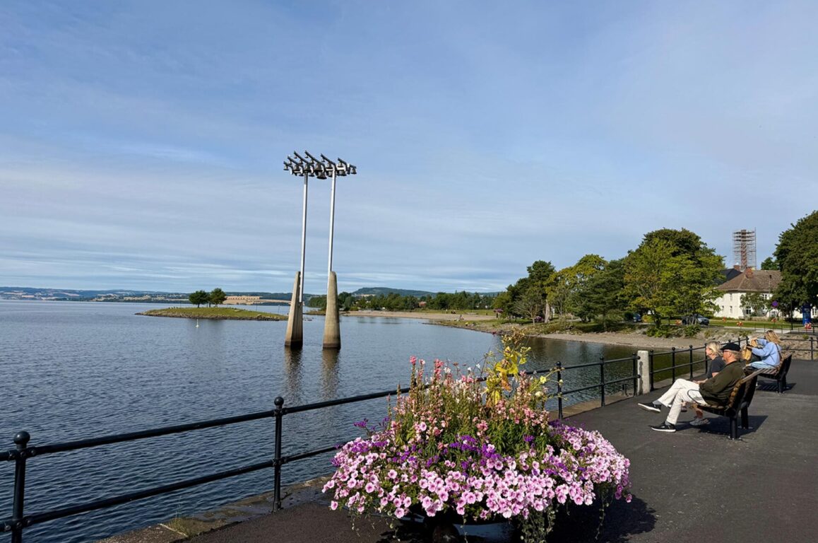 En rolig sjøpromenade med båtstativ i vannet, blomsterkass med rosa og hvite blomster i forgrunn, og benker der tre personer sitter og nyter utsikten mot holmer og trediv.