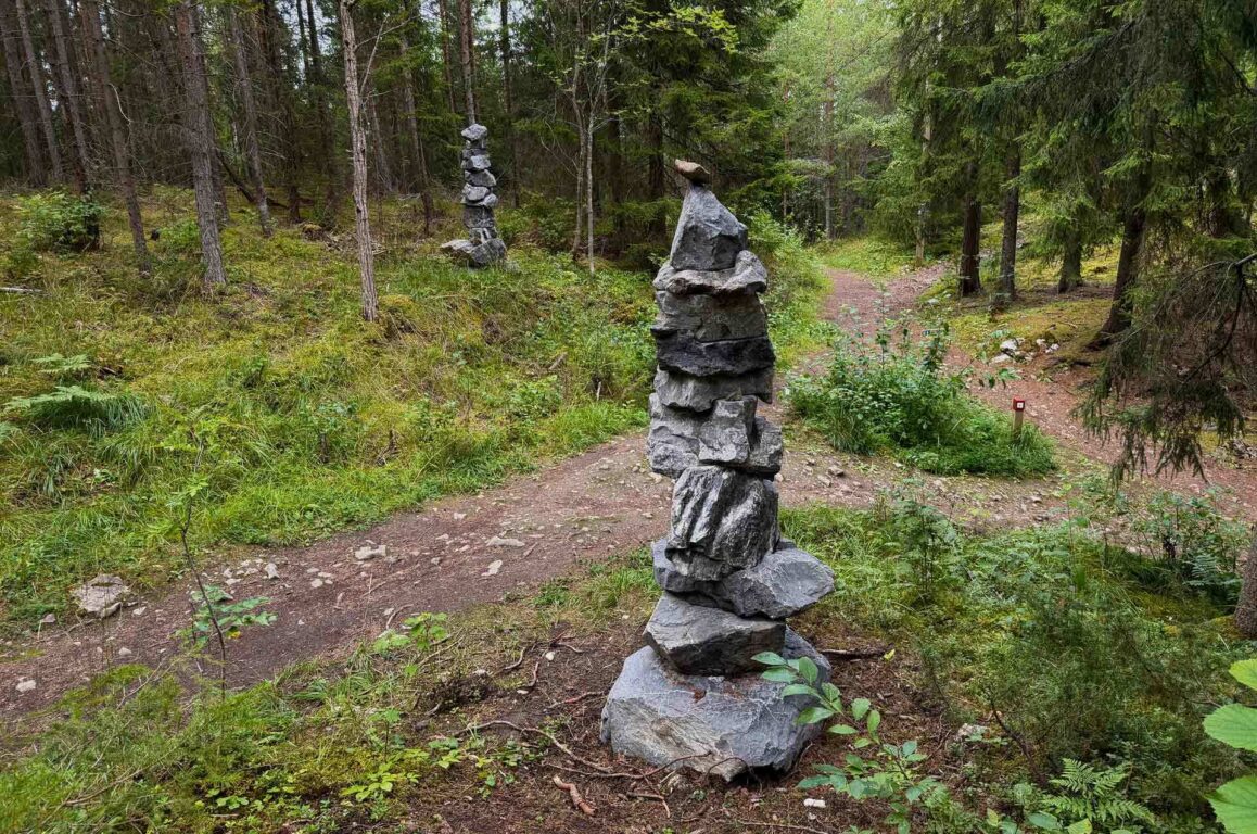 Stier i en skog med flere stable steinfigurer langs stien, høyre og midtre del av bildet viser et større stein-tårn mens andre mindre tårn står i bakgrunnen.