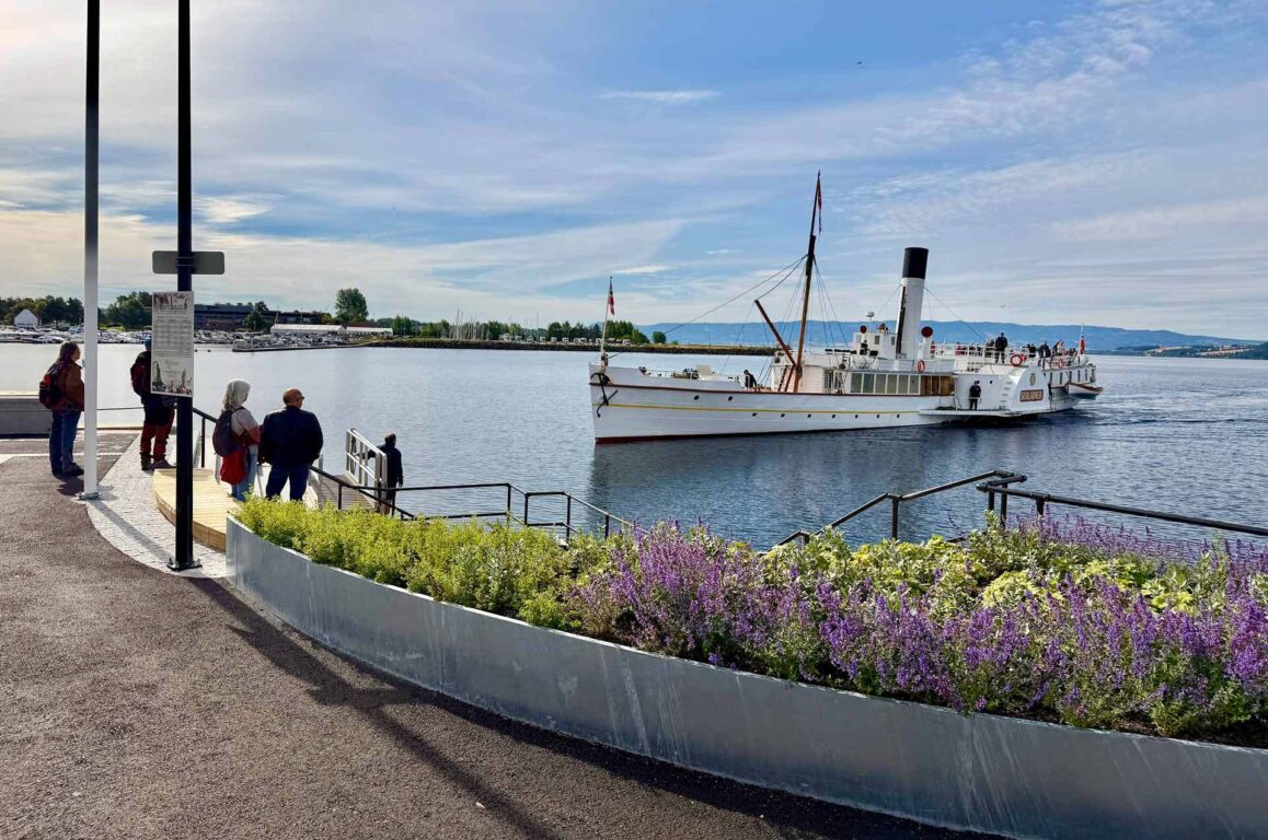 Båtrede ved havnen med en hvit damptogliknende skute og passasjerer ombord; forstikker en rekke fargerike liljer i knust dekorativt bed og en gangvei langs vannet.