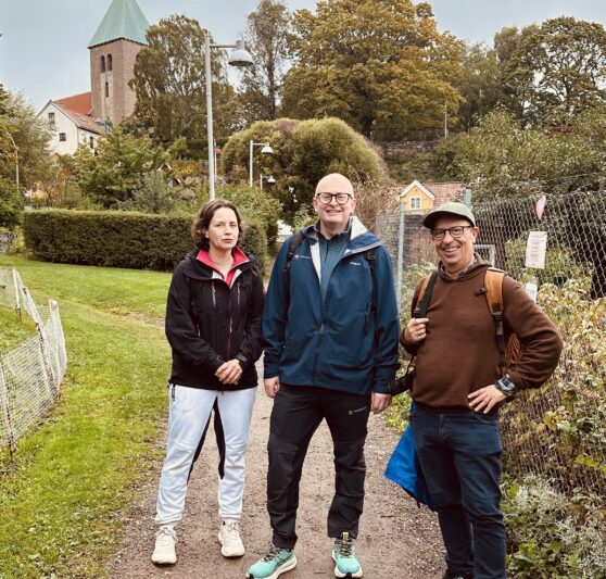 Tre personer står på en grusvei i en parkmessig bydel med trær og bygg i bakgrunnen. De er kledd i utendørsklær og ser mot kamera, en med blå jakke, en med svart jakke og en med bruntgenser og hatt.