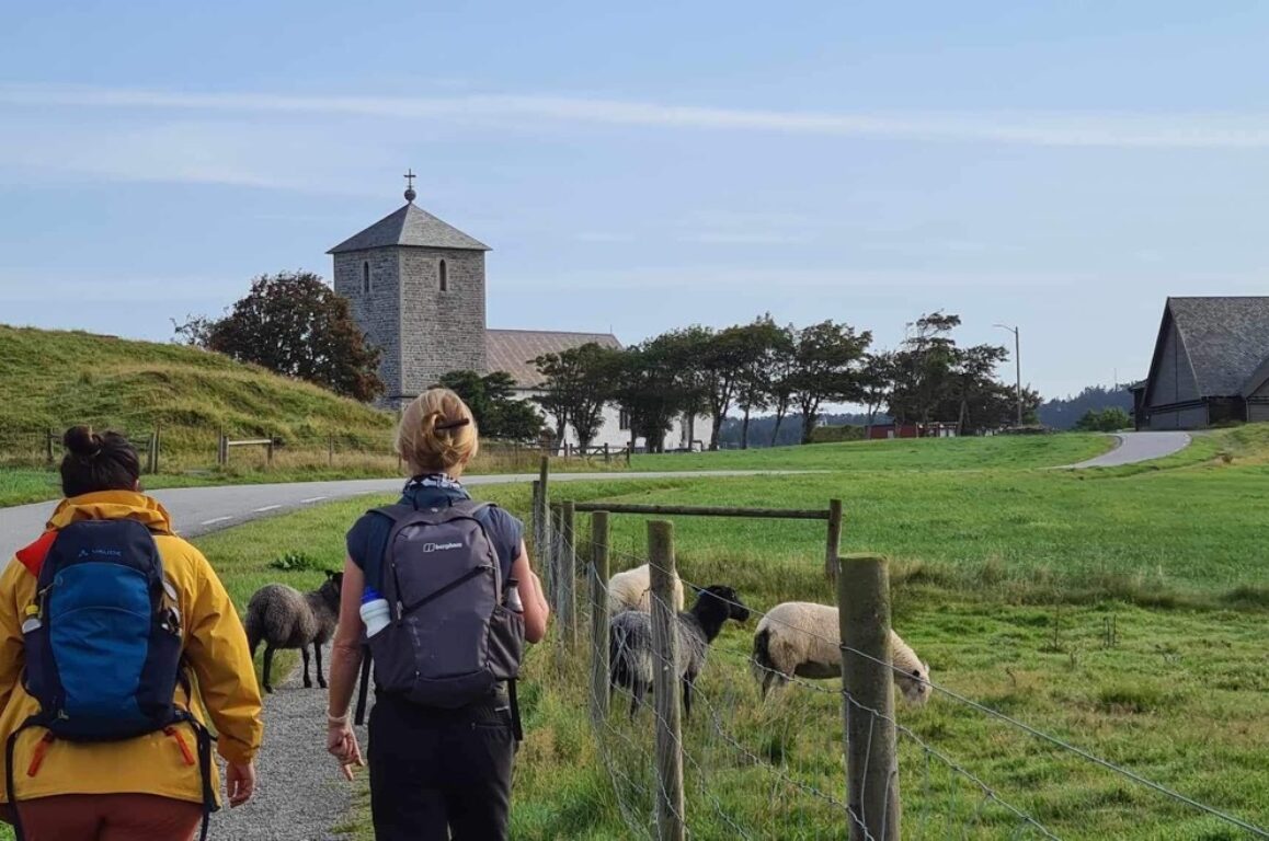 To personer med sekk på ryggen går langs en sti. I bakgrunnen vises en gårdsbygning og en steinkirke.