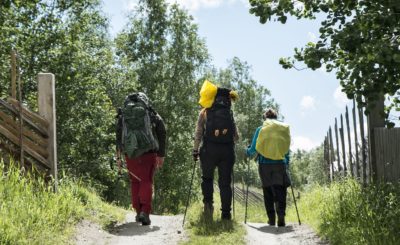 Tre personer går langs en grusvei med sekk på ryggen. Foto