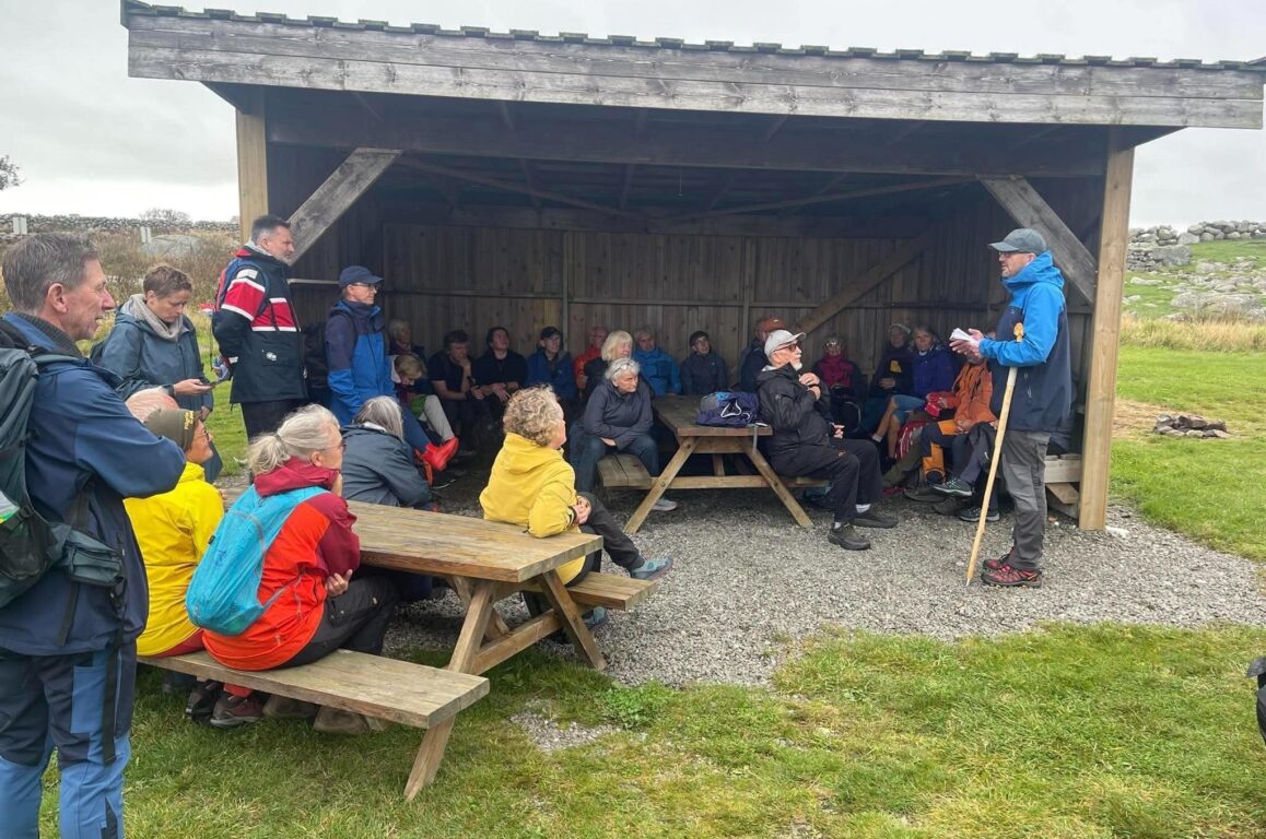 Utendørs gruppemøte i en åpen trek-abbe med bormalt bord, hvor mange personer sitter og står rundt, mens en person står og holder en pinne og snakker til gruppen.