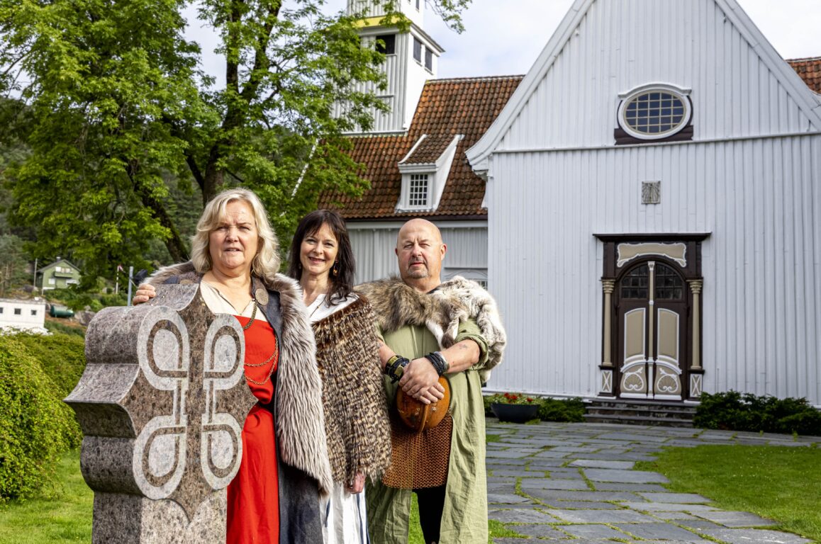 Tre personer bak en stor stein, også kalt milestein. I bakgrunnen: Hvit trekirke.