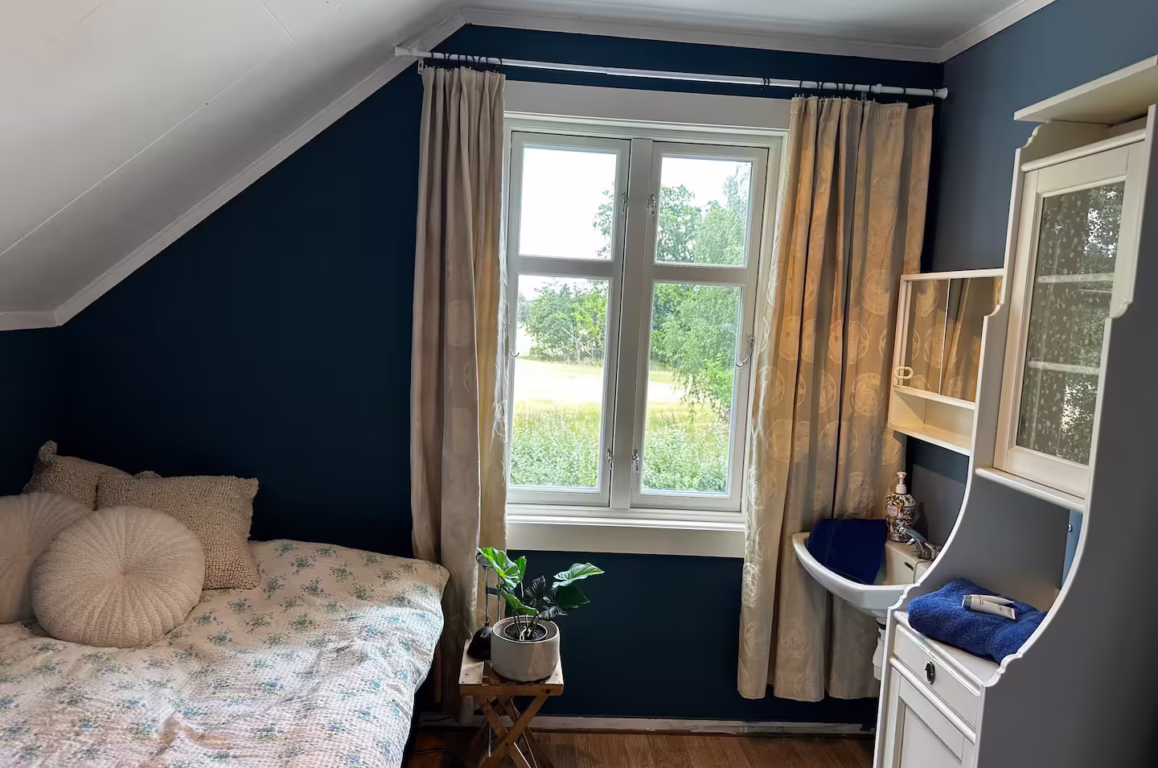 Soverom med mørk blå vegger, skjevvånn skråtak, svalt vindu med beige gardiner og sengetøy med blomster. Bord ved vinduet med plante og soveligner.