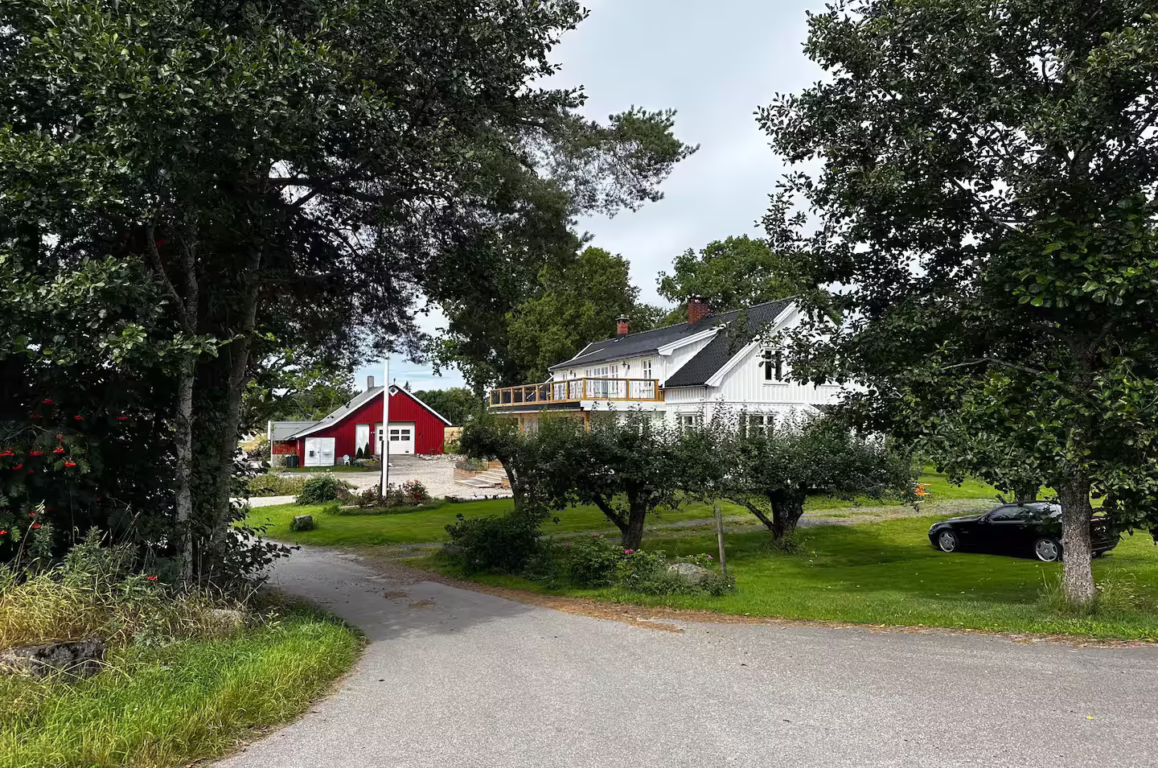 Frittliggende gårdsområde med hvit hus og terrasse omgitt av grøntområde, rødt bygg i bakgrunnen, bil på gresset og veiskråning i forgrunnen.