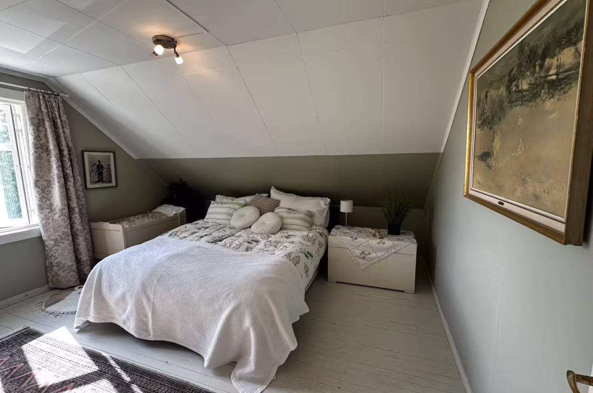 soverom med skråtak og lyst grønnmalt vegger, seng med hvit sengetøy og putepar, nattbord med lampe, vindu med patterned gardiner, sidestoff og malerisk landskapsbilde på vegg.