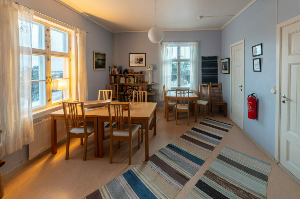 Oppholdsrom med to spisestuebord, hylle med lesestoff, filleryer på gulvet og utsikt over øy og hav.